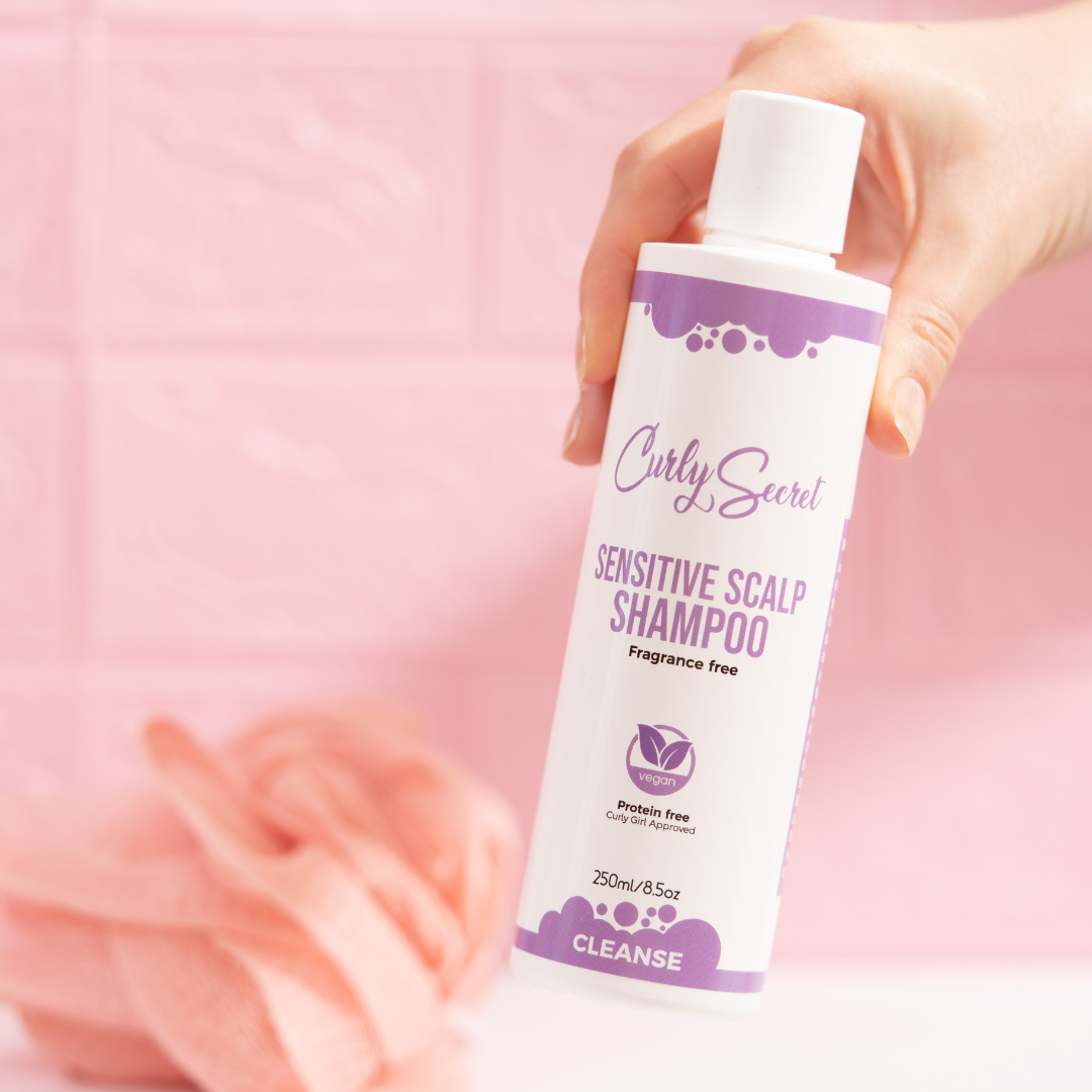 Hand holding Curly Secret Sensitive Scalp Shampoo bottle, pink background, hair care product