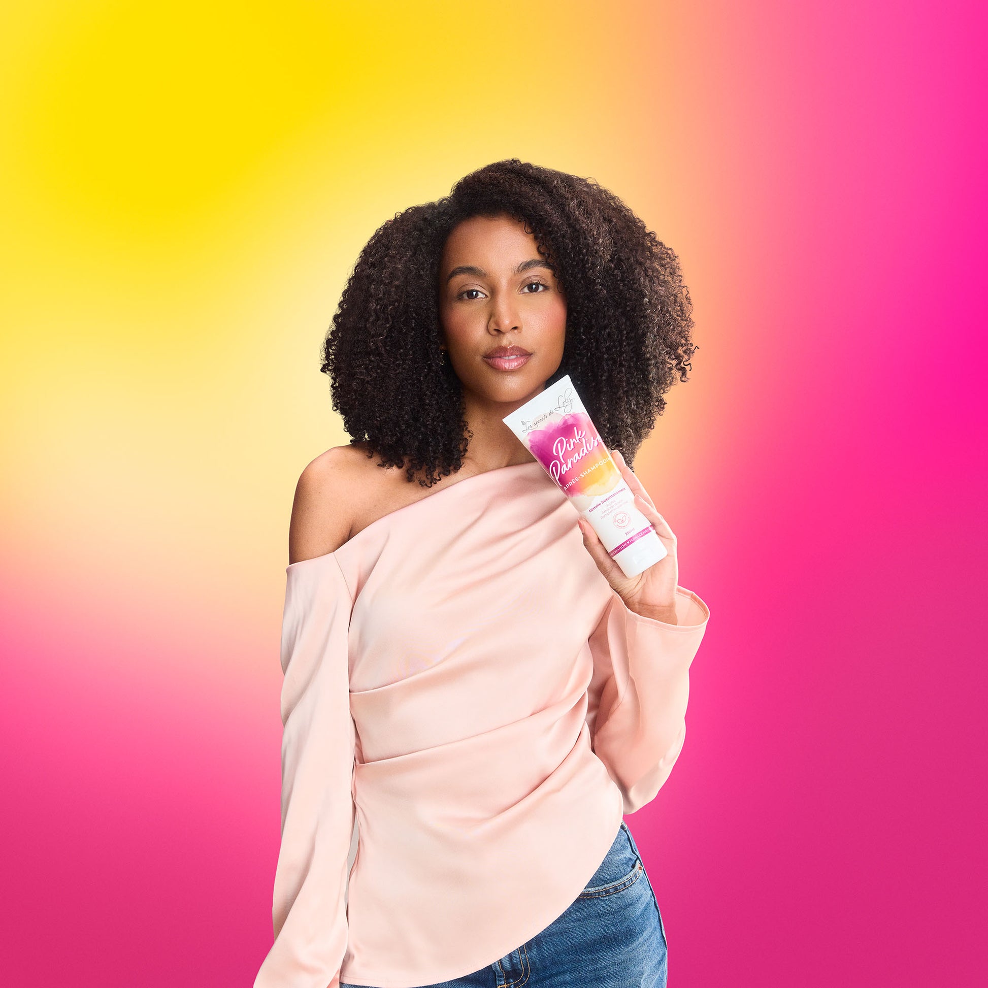 Woman with natural curly hair holding hair care product against vibrant pink and yellow background