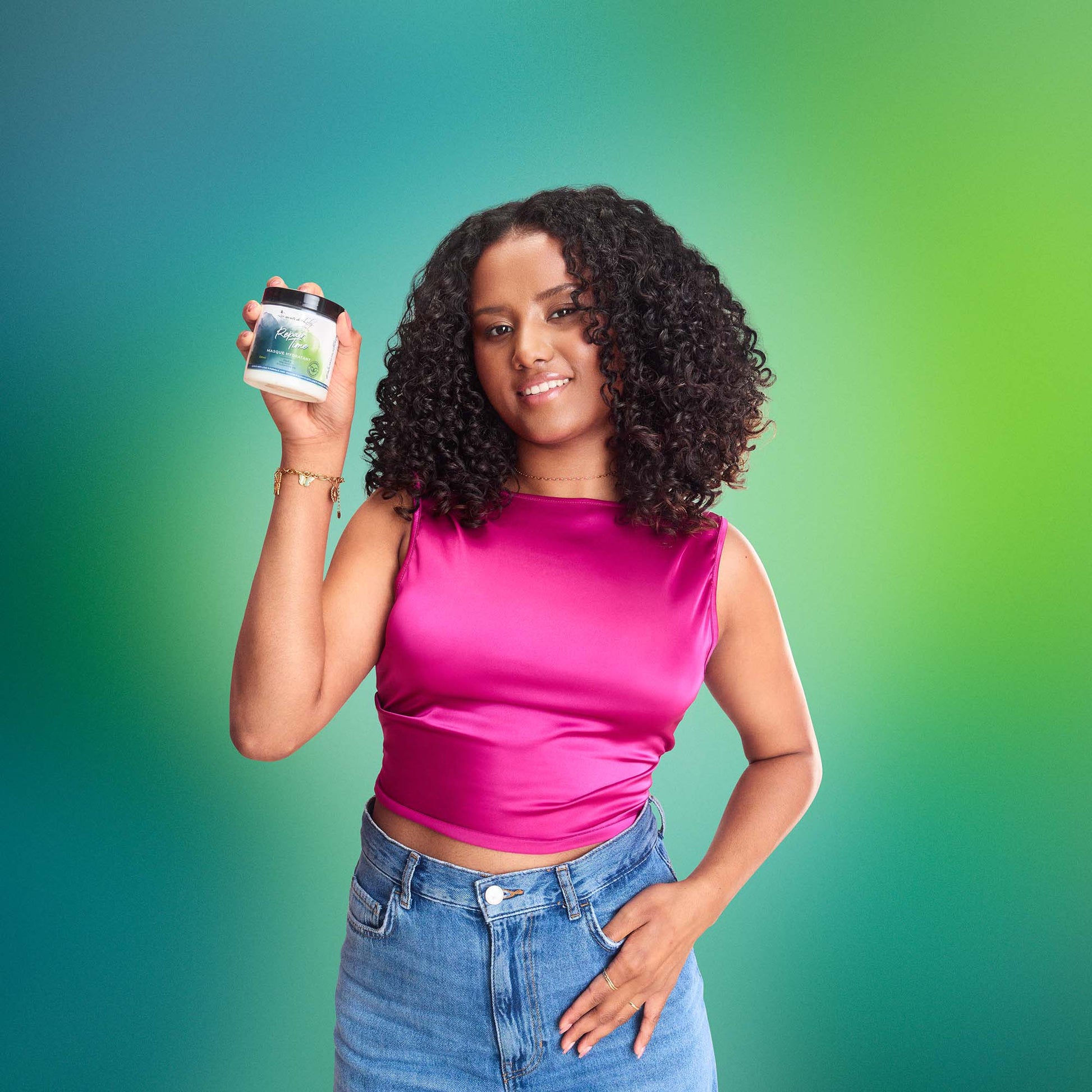 Smiling woman with curly hair holding natural hair care product against green background